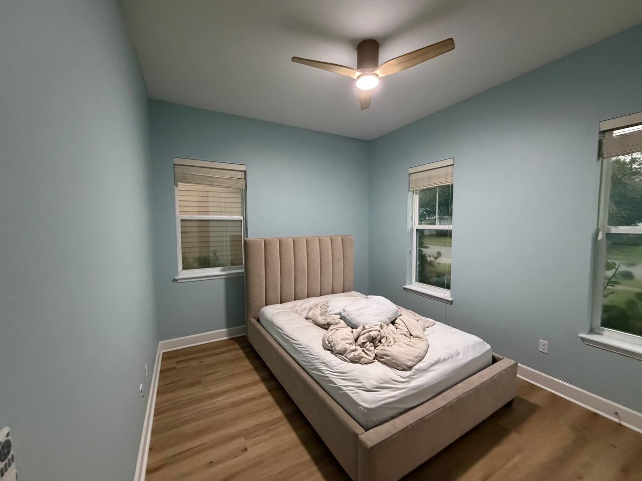 After: the finished bedroom from the doorway, soft blue-green walls, ceiling fan and clear wood floor.