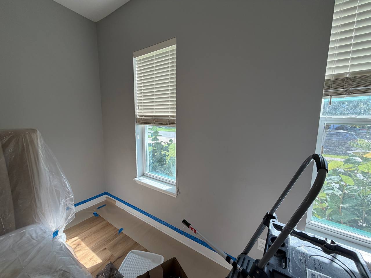 Before: the same bedroom corner in grey, baseboards masked with blue tape and spray equipment on the floor.
