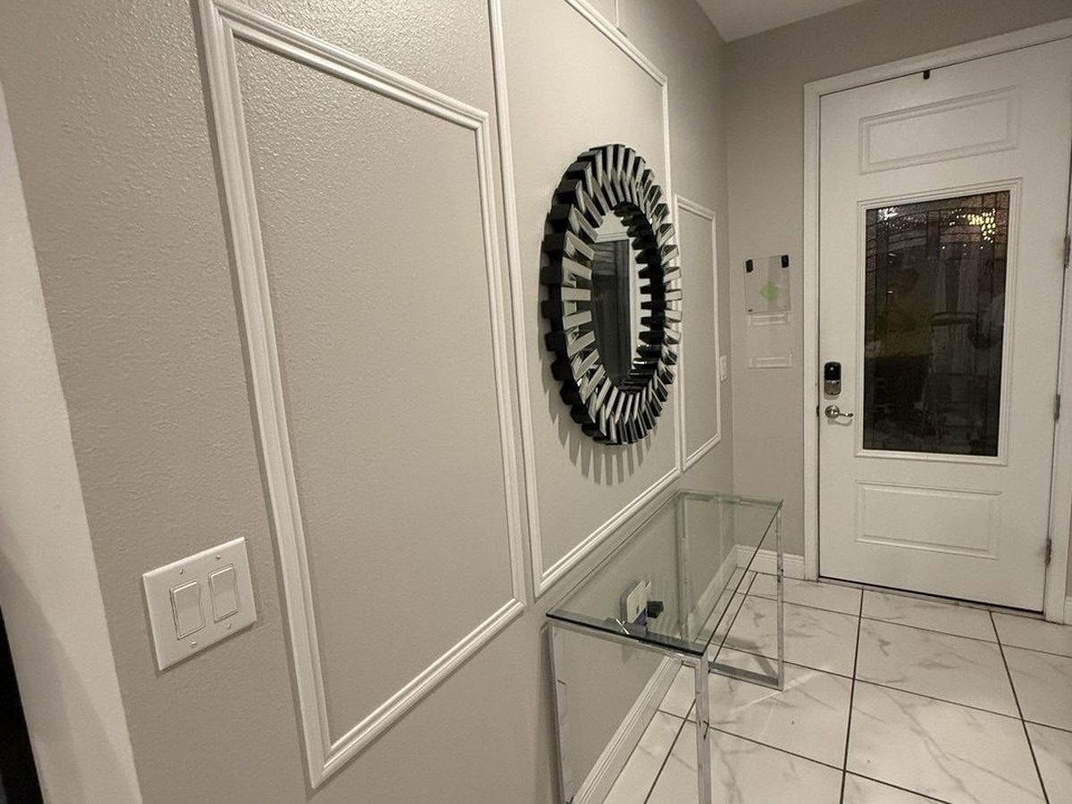 Home entryway with white picture-frame trim panels applied to the wall, a round sunburst mirror and a glass console table.